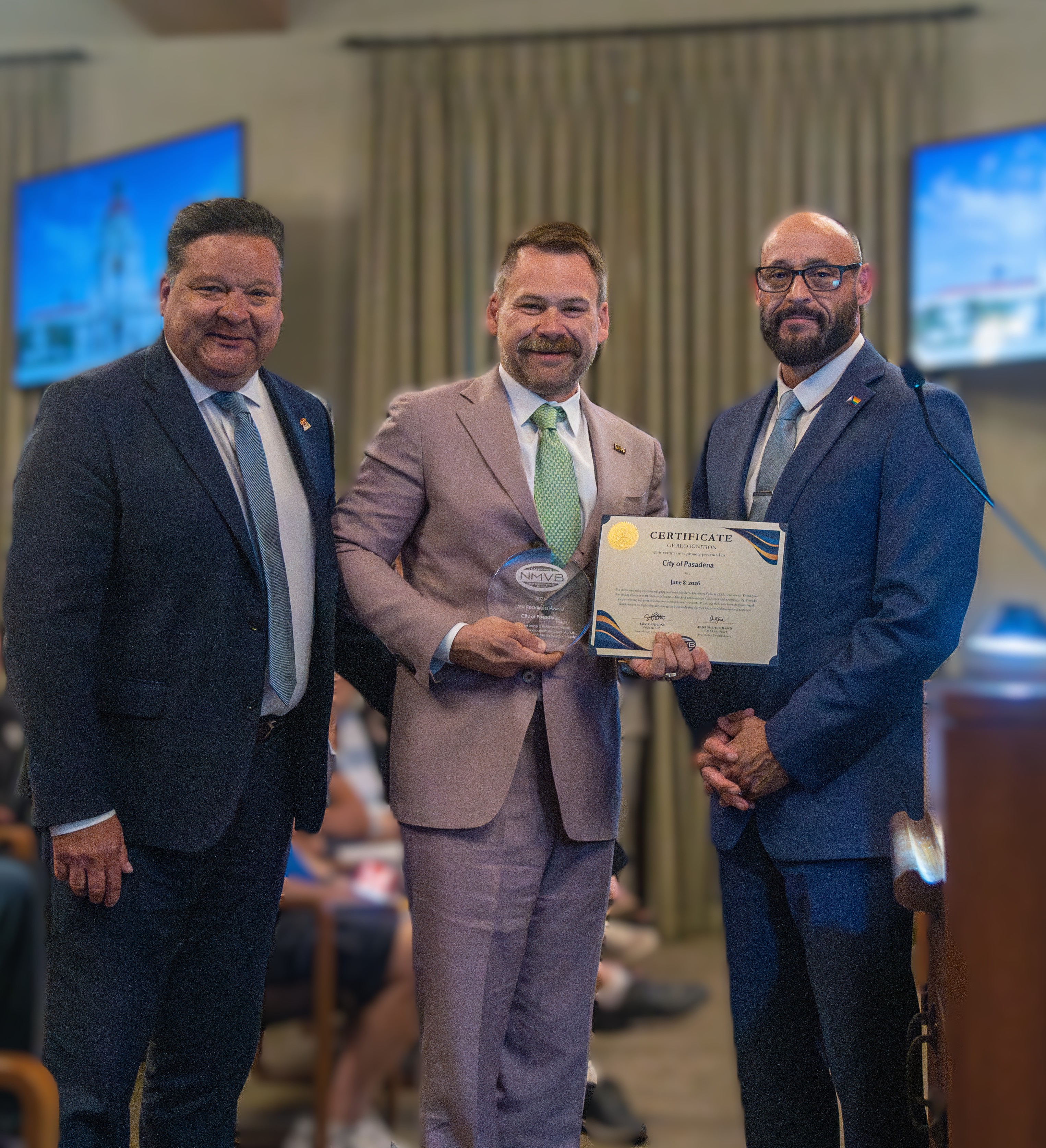 NMVB Board President Jacob Stevens presenting a ZEV Readiness Award certificate and glass award to two City of Pasadena representatives at a council meeting.