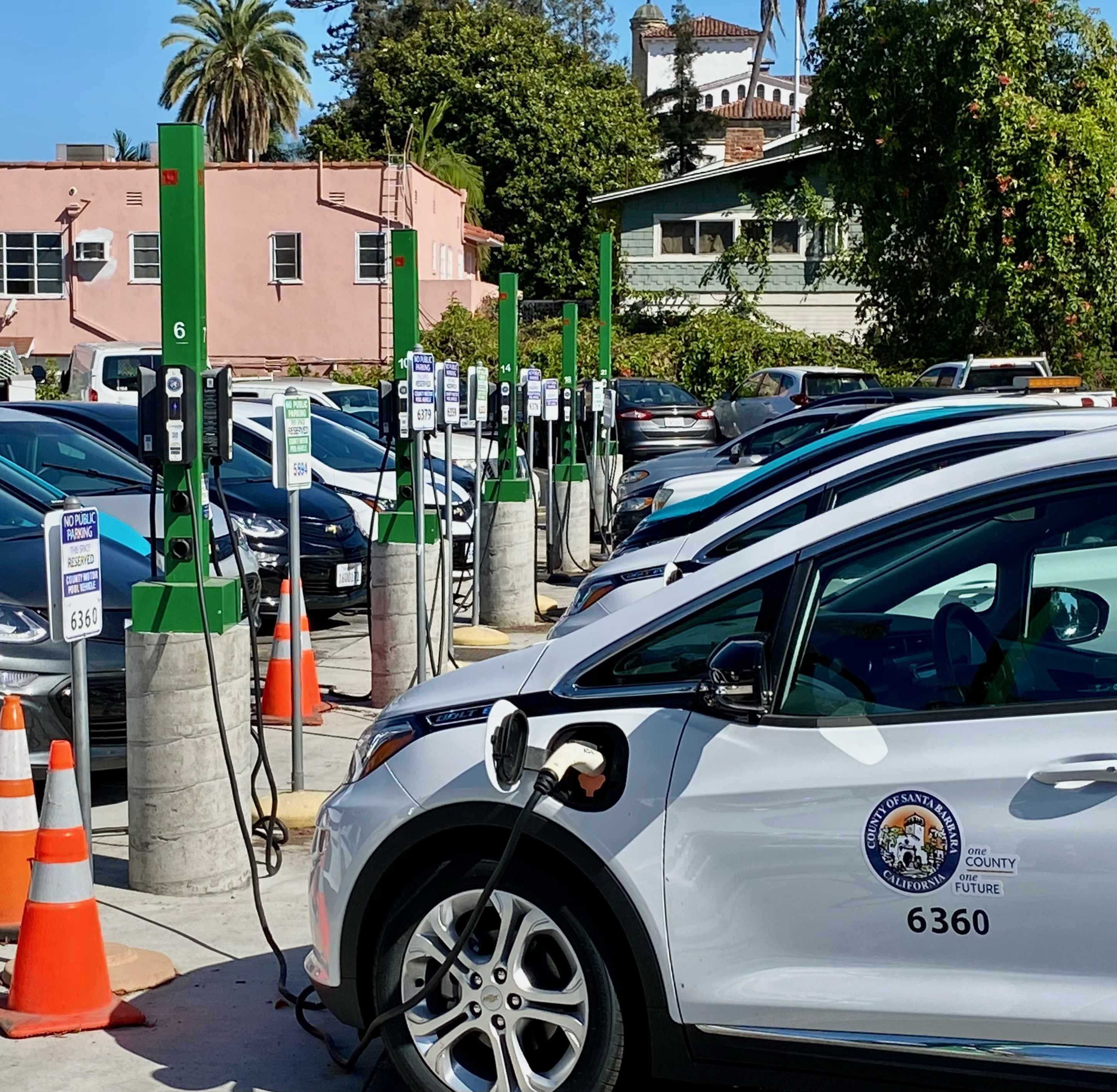 A row of County of Santa Barbara fleet electric vehicles plugged into green charging stations in a county parking lot.